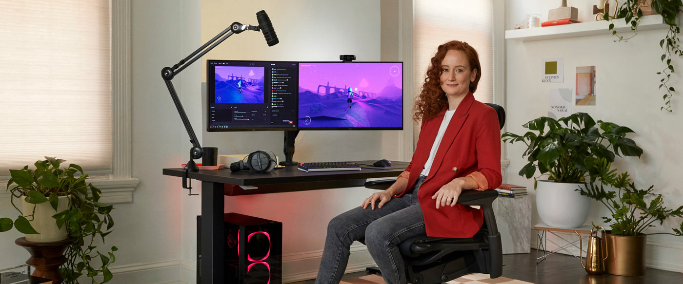 Foto van Dr. Lindsey Migliore zittend in een Embody gamingstoel, met de camera aan, met een Nevi-gamingtafel in zwart en monitorarmen van Ollin