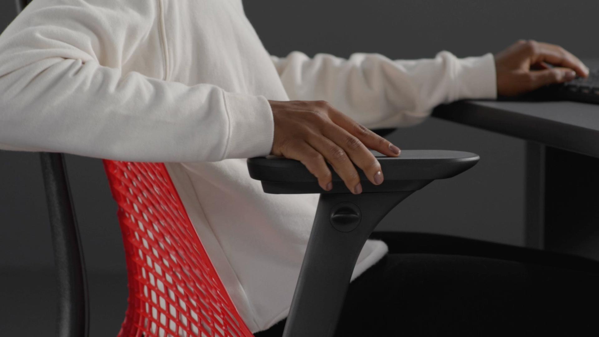 Een video van een persoon die in een rood-zwarte Sayl-gamingstoel van Herman Miller zit en naast de stoel reikt om de armleuningen te verstellen.