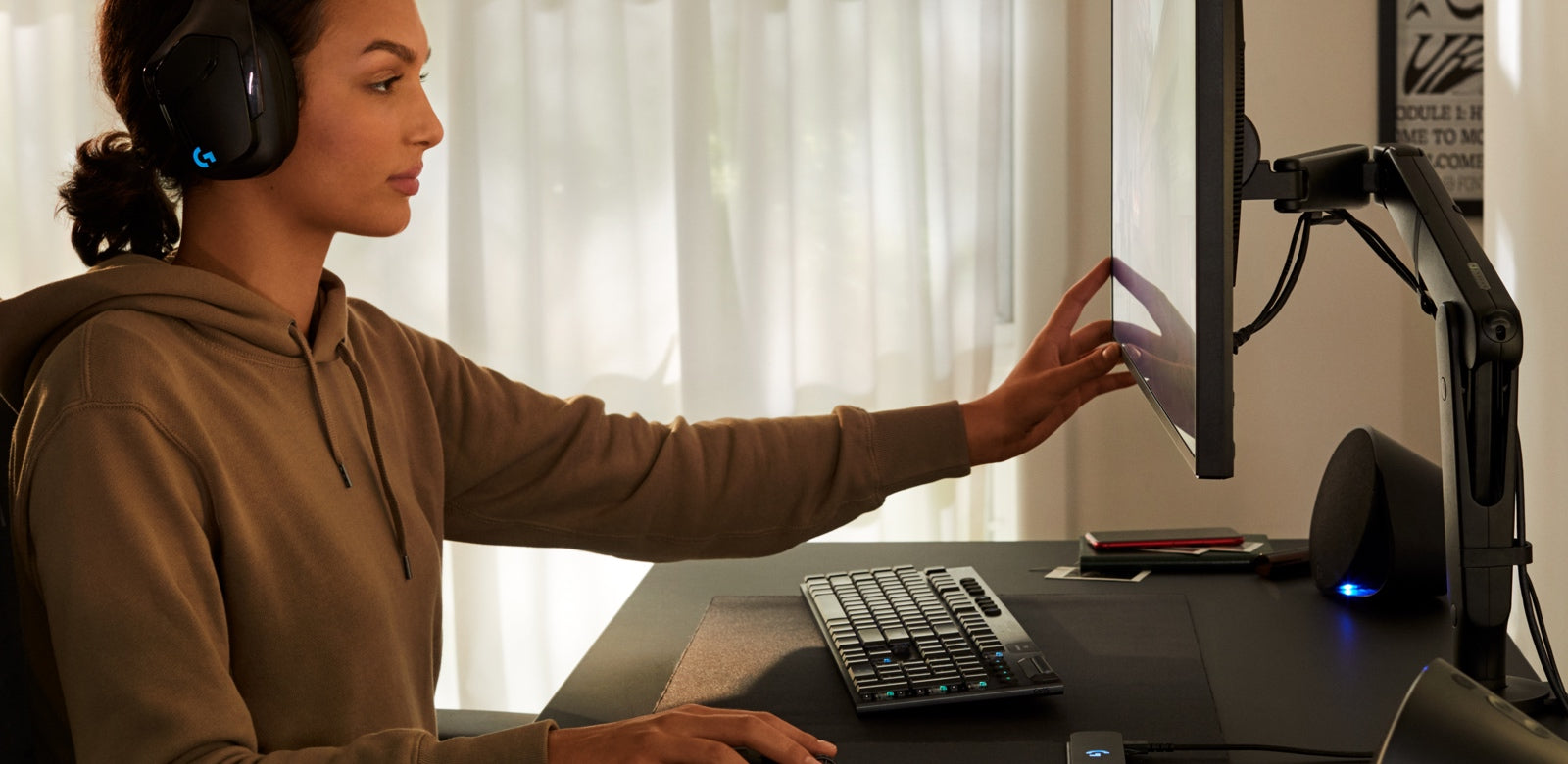 Een vrouw zit aan een zwart Nevi-gamingbureau met een zwarte Ollin-gamingmonitorarm van Herman Miller. Ze draagt een Logitech-koptelefoon en beweegt het computerscherm met één van haar handen.