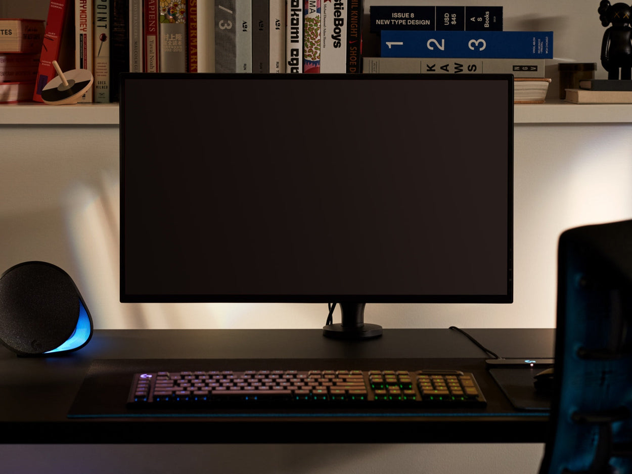 Vooraanzicht van een zwarte Ollin-monitorarm met een computerscherm, bevestigd aan een zwart Nevi-gamingbureau van Herman Miller. Geplaatst in een gameroom of thuiskantoor, met een boekenplank, toetsenbord en muis.