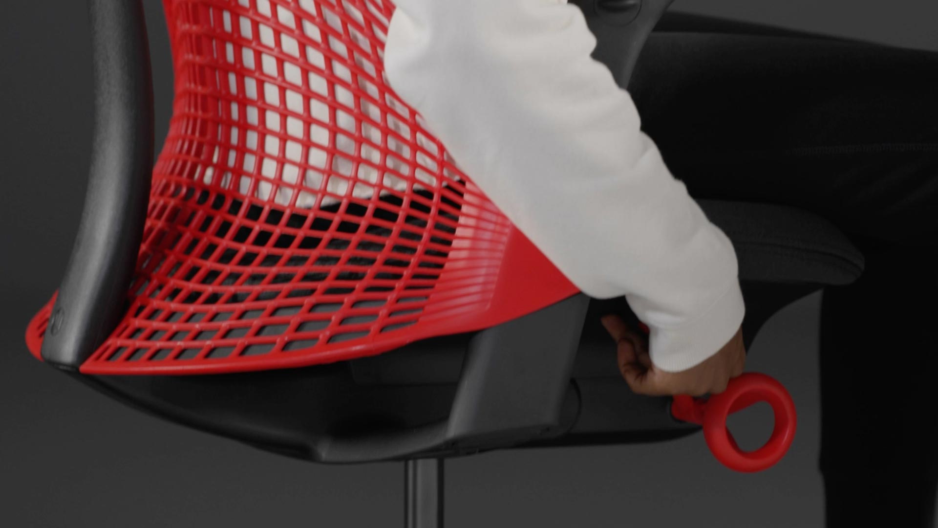 Een video van een persoon die in een rood-zwarte Sayl-gamingstoel van Herman Miller zit en naast de stoel reikt om de zithoogte aan te passen.