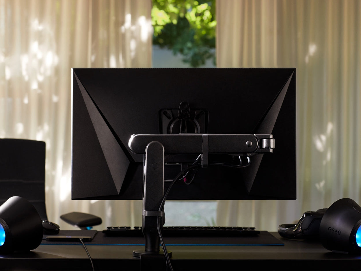 Achteraanzicht van een zwarte Ollin-monitorarm met een computerscherm, bevestigd aan een zwart Nevi-gamingbureau van Herman Miller. Geplaatst in een gameroom of thuiskantoor, met lampen, toetsenbord en muis.
