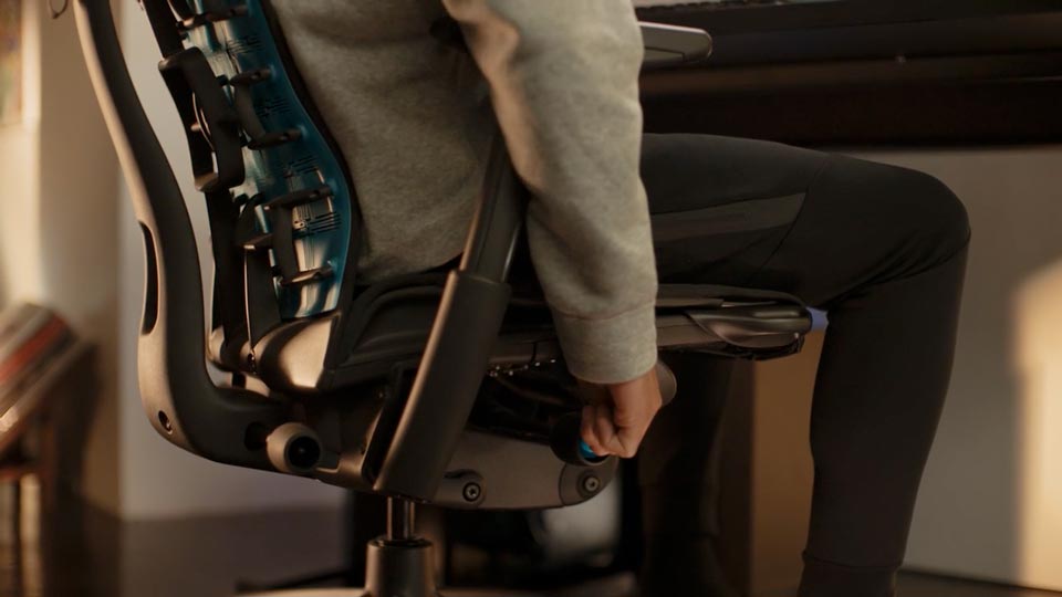 Een video van een persoon die in een zwart-blauwe Embody-gamingstoel van Herman Miller zit en naast de stoel reikt om de zithoogte aan te passen.