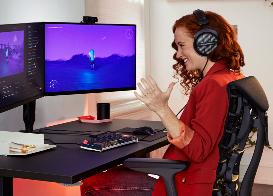 Foto van Dr. Lindsey Migliore die glimlachend een computerspel speelt in een Embody cyan gamingstoel met een Nevi gamingtafel in zwart