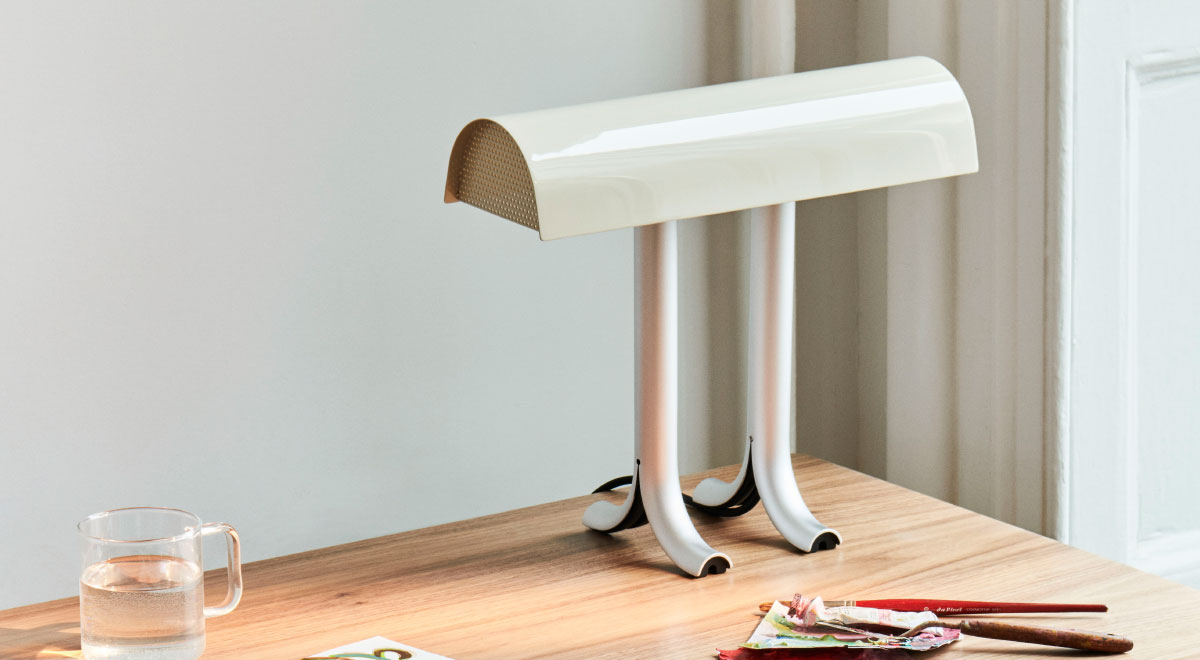 Een crèmekleurige en zilveren Anagram-tafellamp van HAY, geplaatst in de achterste hoek van een houten bureau.