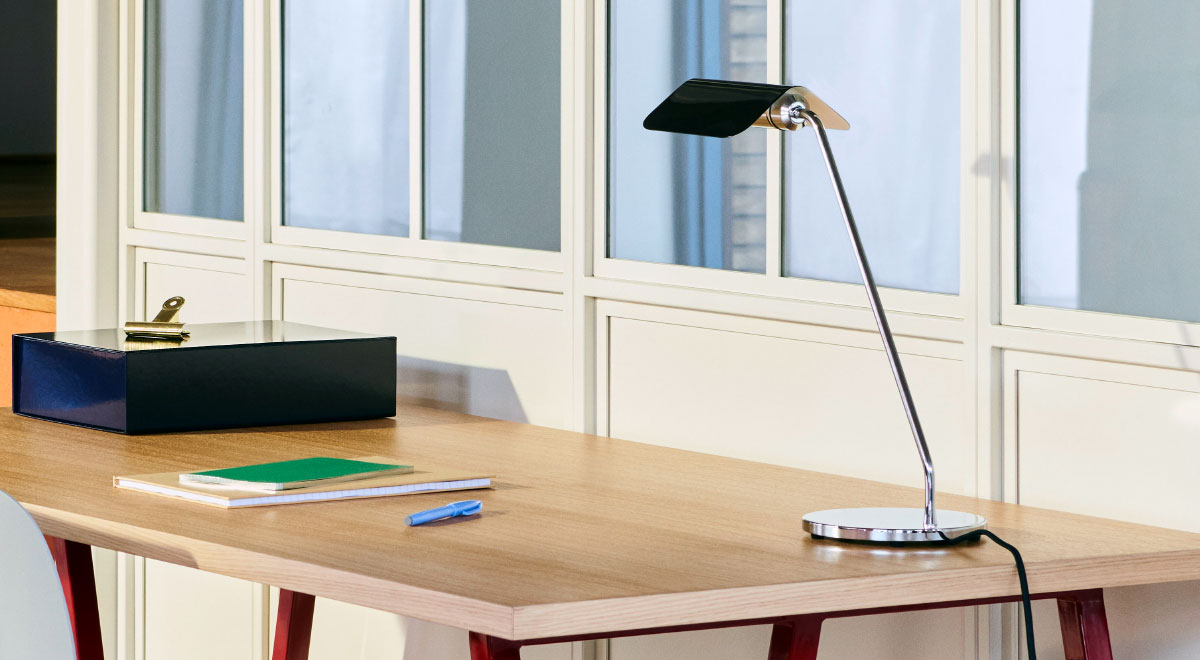 Een zwart-zilveren Apex-bureaulamp van HAY, geplaatst in de achterste hoek van een houten bureau.
