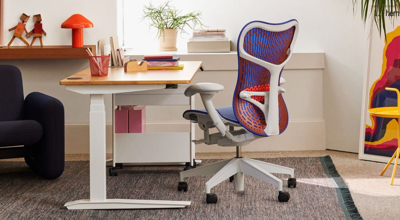 Een blauwe, oranje en witte Mirra 2-bureaustoel van Herman Miller, geplaatst naast een Jarvis-sta-bureau. Geplaatst in een thuiskantooromgeving met een blauwe Wilkes-bank, blauw Maharam-tapijt, gele DLM-bijzettafel en rode HAY Pao-tafellamp.