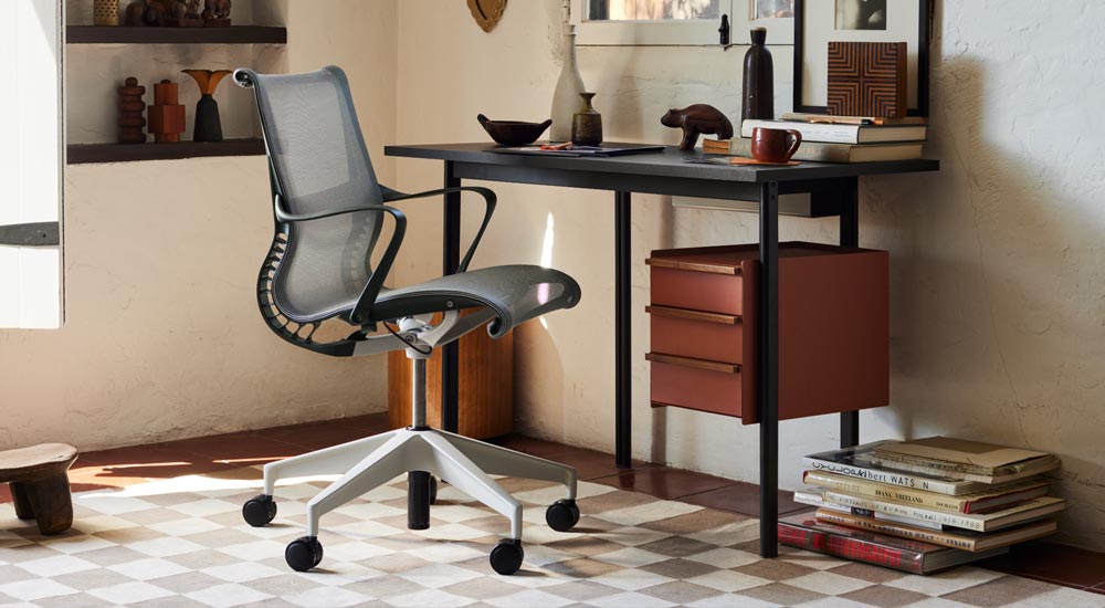 Een zwart Mode-bureau van Herman Miller, met terracotta opbergruimte met walnoothouten handgrepen, staat voor een grijze Setu-bureaustoel van Herman Miller. Geplaatst in een goed ingerichte thuiskantoor met planken, boeken en decoratieve woonaccessoires.