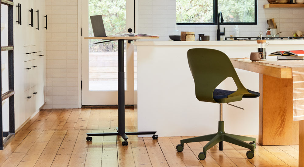 Een zwarte en eikenhouten Passport-tafel van Herman Miller, geplaatst naast een wit kookeiland, met een laptop en een boek. Op de afbeelding is ook een groene en zwarte Zeph-stoel van Herman Miller te zien, geplaatst aan een houten eettafel.