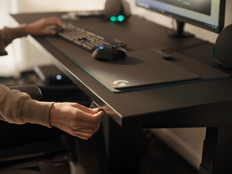 Een video van een persoon die aan een zwart Nevi-gamingbureau van Herman Miller zit en op een knop drukt om het bureaublad te verlagen.
