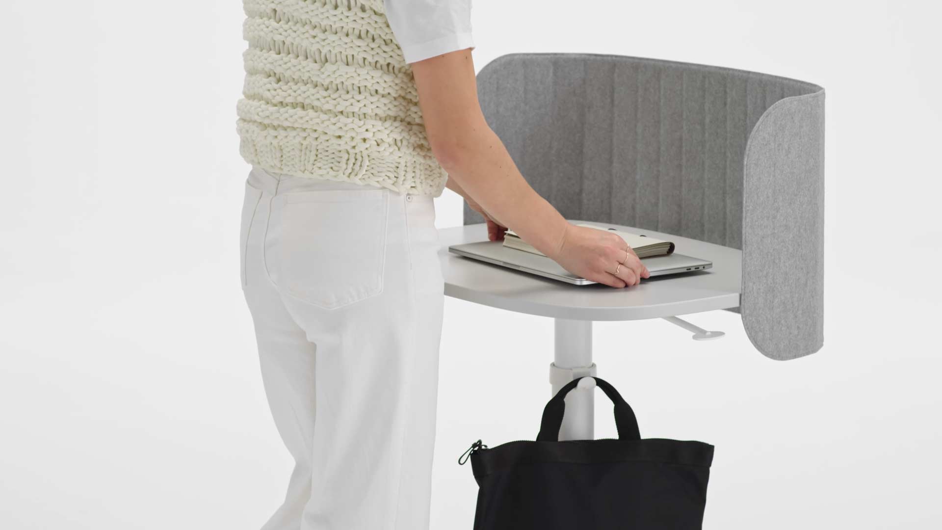 Een video van een vrouw die een witte Passport-werktafel gebruikt. Ze loopt naar het bureau, hangt haar tas aan de tassenhaak, plaatst haar laptop op het tafelblad en zet het bureau vervolgens op de juiste hoogte.
