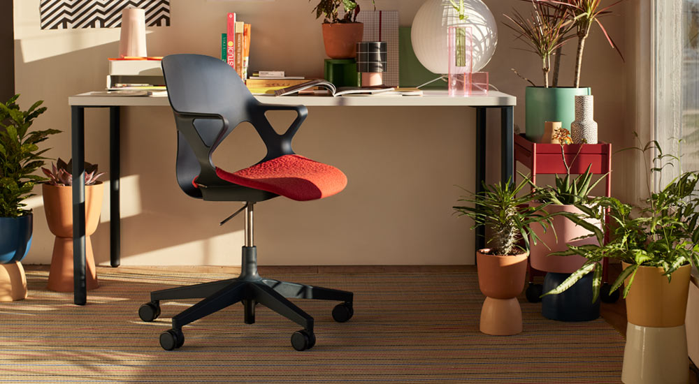 Een zwarte Zeph-bureaustoel met een rode zitkussen staat voor een Herman Miller OE1-bureau. Geplaatst in een thuiskantooromgeving, met een rode OE1-trolley en een selectie kamerplanten in kleurrijke potten.