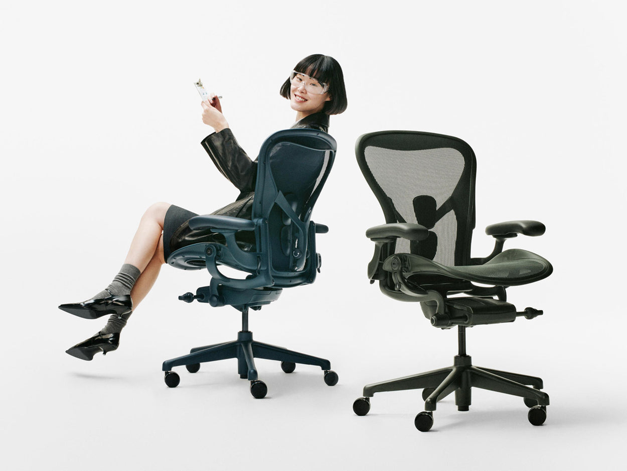 In een witte studio zat een vrouw op een blauwe Herman Miller Aeron ergonomische bureaustoel van het model Nighfall, naast een andere Aeron in de kleur Jasper Green.