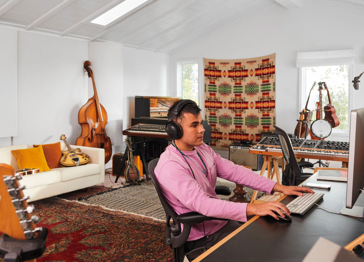 Rostam, muzikant, producer en componist, zit in een Aeron-bureaustoel in grafiet achter een computer met een koptelefoon op en muziekinstrumenten op de achtergrond.