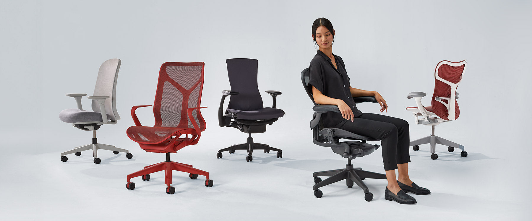 Foto van Herman Miller-stoelen op een rij, waaronder Verus mineral white & grey, Cosm mid back in canyon red, Embody black, Aeron graphite met een dame erin en Mirra 2 in cordovan red