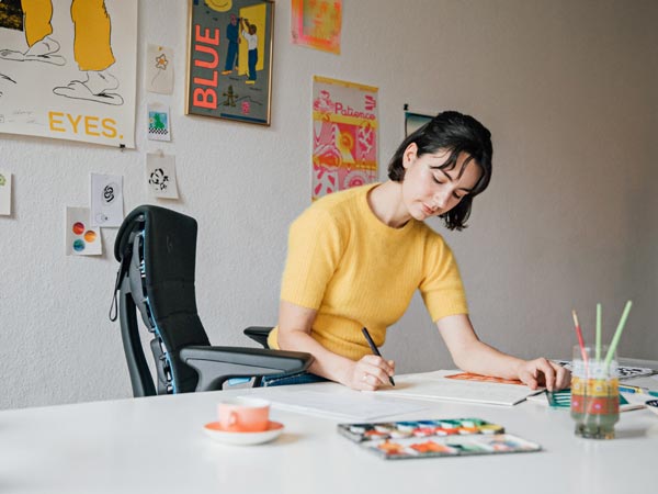 Illustrator, schilder en auteur Molly Rose Dyson aan het werk in haar studio, zittend in een Embody-gamingstoel in zwart en cyaanblauw