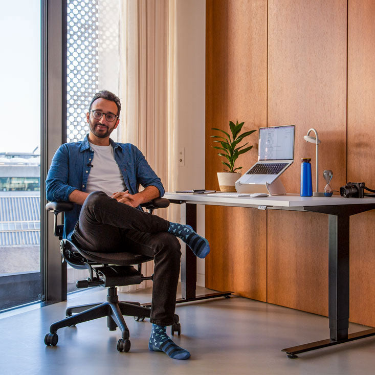 Foto van Ali Abdaal, podcaster, zittend in een Herman Miller Aeron-bureaustoel in grafiet voor een Nevi-sta-zitbureau met zwarte poten en een wit blad, met een laptop in een Oripura-laptopstandaard