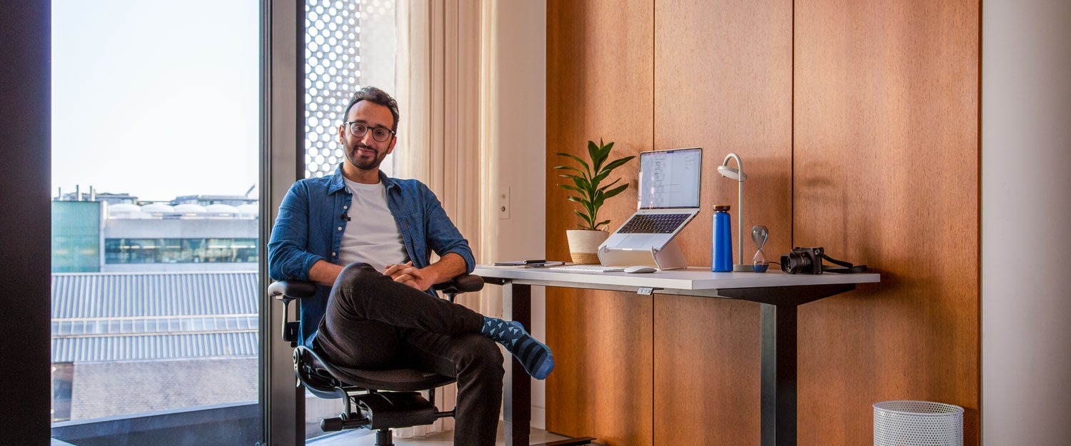 Foto van Ali Abdaal, podcaster, zittend in een Herman Miller Aeron-bureaustoel in grafiet voor een Nevi zit-sta-bureau met zwarte poten en een wit blad, met een laptop in een Oripura-laptopstandaard