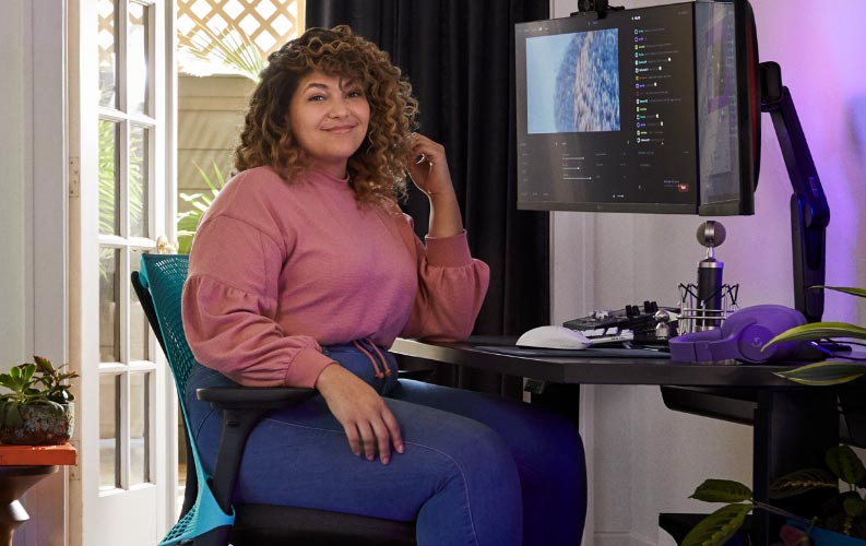 Professionele streamer en stemacteur Friskk zittend op een Herman Miller Sayl-gamingstoel in diepblauw en zwart, aan een zwarte Nevi-sta- en zit-gamingtafel met monitor op een Oliin-monitorarm.