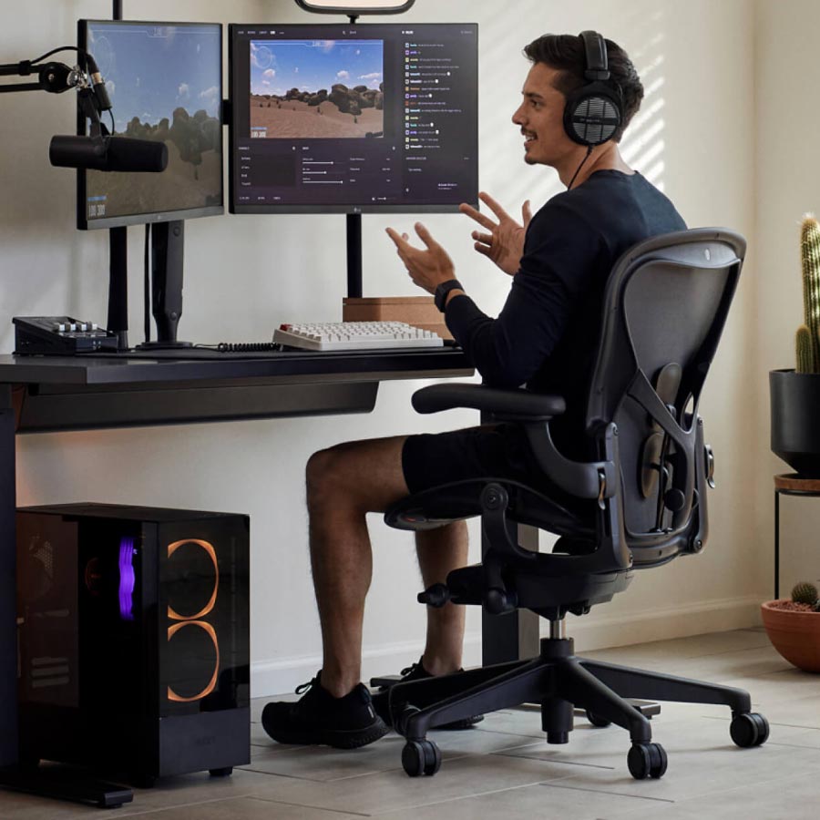 Gamer en streamer Xaryu in een gamingopstelling, zittend op een Herman Miller Aeron-gamingstoel met een zwart Nevi-sta-zit-gamingbureau en twee monitoren op Ollin-monitorarmen