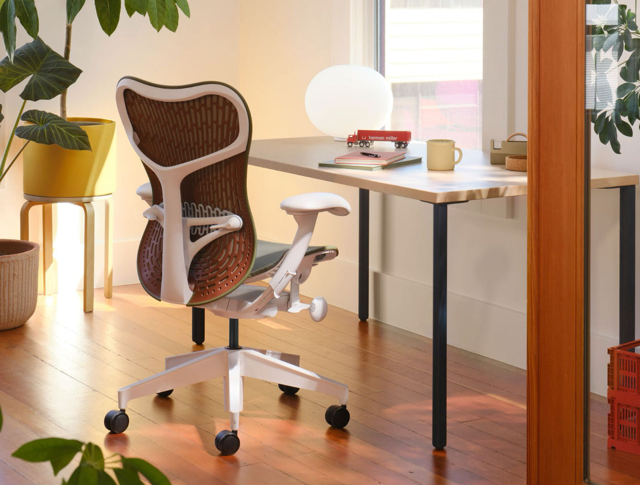 Een olijfgroene en witte Mirra 2-bureaustoel met een kastanjebruine rugleuning, geplaatst voor een Herman Miller OE1-bureau. Geplaatst in een thuiskantooromgeving, met massief houten vloeren, witte muren en warme, gezellige verlichting.