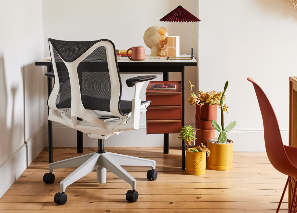 Een blauw-witte Cosm-bureaustoel van Herman Miller, geplaatst voor een zwart en terracotta Mode-bureau van Herman Miller. Geplaatst in een thuiskantoor met houten vloeren en witte muren, aangevuld met een Matin-tafellamp en kamerplanten.