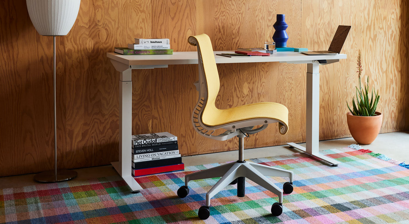 Een gele Setu-bureaustoel van Herman Miller, bekleed met Mimosa Meld-stof, geplaatst voor een wit Nevi-sta-bureau op een kleurrijk geruit tapijt.