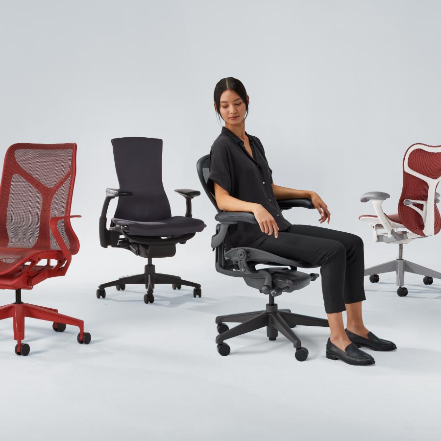 Vier Herman Miller-bureaustoelen op een rij, waaronder een Cosm met middenhoge rugleuning in canyonrood, een Embody in zwart, een Aeron in grafiet met een vrouw erop en tot slot een Mirra 2 met een wit frame en een rode zitting en rugleuning