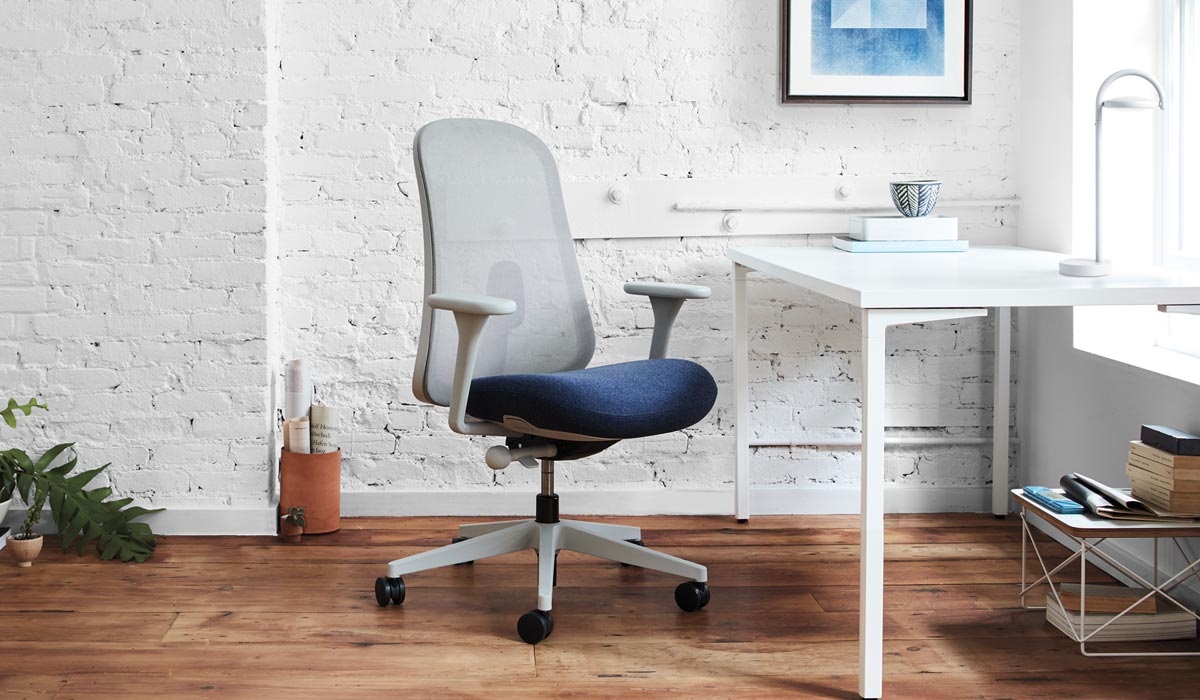 Een grijze en blauwe Lino-bureaustoel van Herman Miller staat voor een wit bureau. De binnenruimte heeft een witte bakstenen muur, houten vloer en een witte Marselis-tafellamp op het bureau.