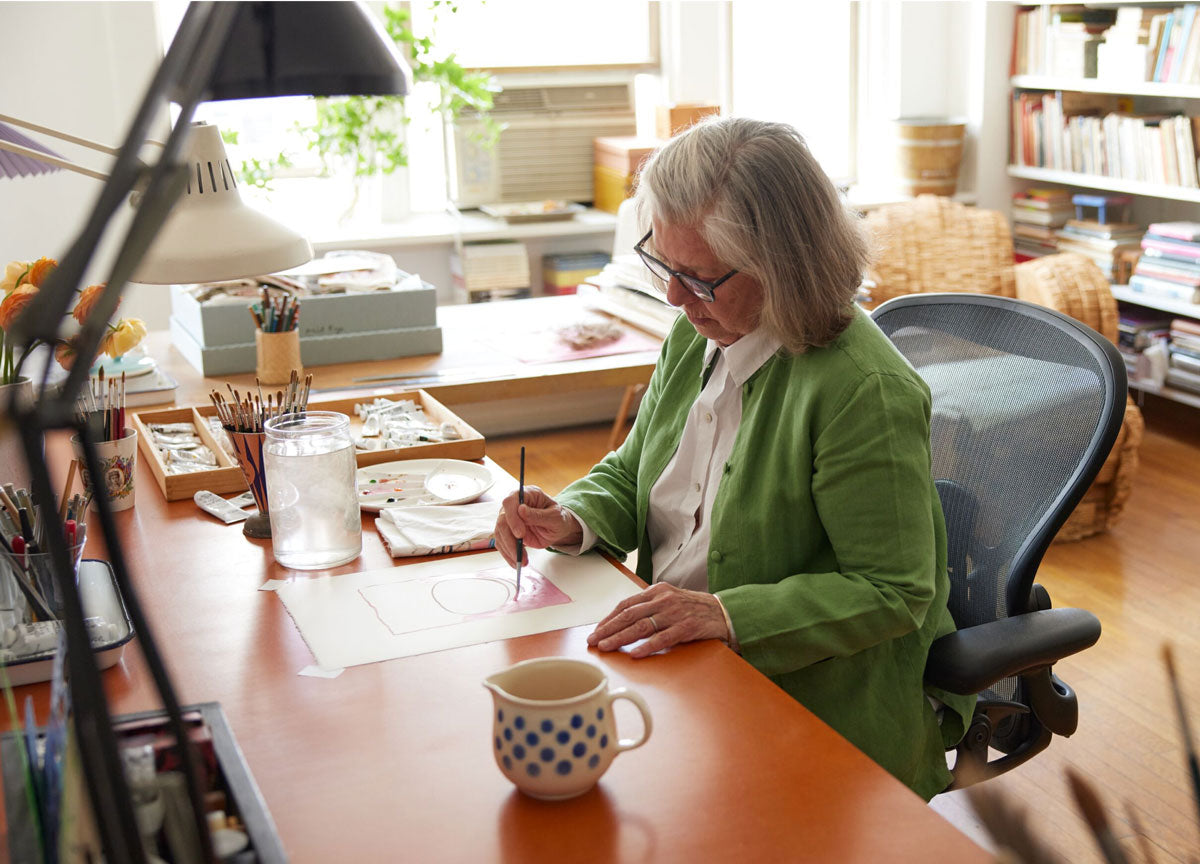 Kunstenares Maira Kalman werkt aan een schilderij in een lichte studioruimte vanaf haar Onyx Herman Miller Aeron.