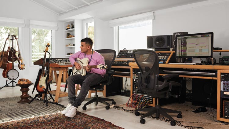 Foto van Rostam, muzikant, producer en componist, in zijn muziekstudio terwijl hij gitaar speelt in een Aeron-bureaustoel in grafiet