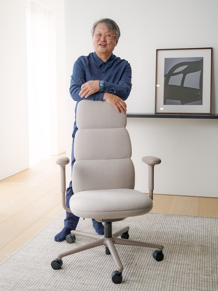 De ontwerper, Naoto Fukasawa, leunt op een Herman Miller Asari-bureaustoel. De setting heeft een strakke vloer en muur op de achtergrond.