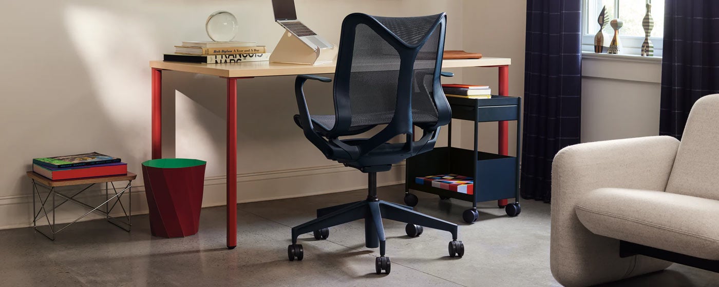 Een blauwe Cosm-bureaustoel van Herman Miller, geplaatst voor een rood en essenhouten OE1-bureau van Herman Miller. Geplaatst in een thuiskantoor, aangevuld met een blauwe OE1-trolley, een witte Oripura-laptopstandaard en een rode HAY Paper-prullenbak.