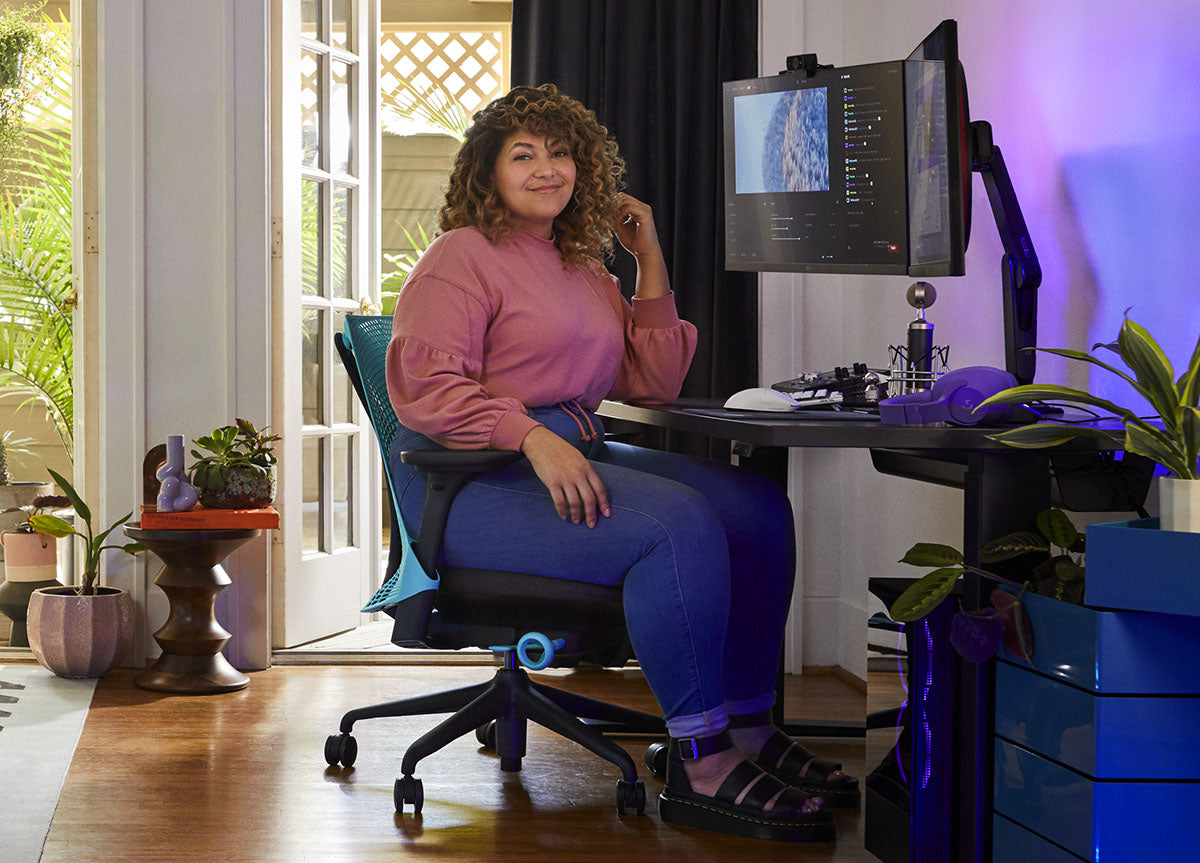 Een vrouw zit in een blauw-zwarte Sayl-gamingstoel van Herman Miller, achter een zwart Nevi-bureau met een zwarte Ollin-monitorarm en -scherm. De foto is genomen in een binnenruimte met houten vloeren, witte muren en kamerplanten.