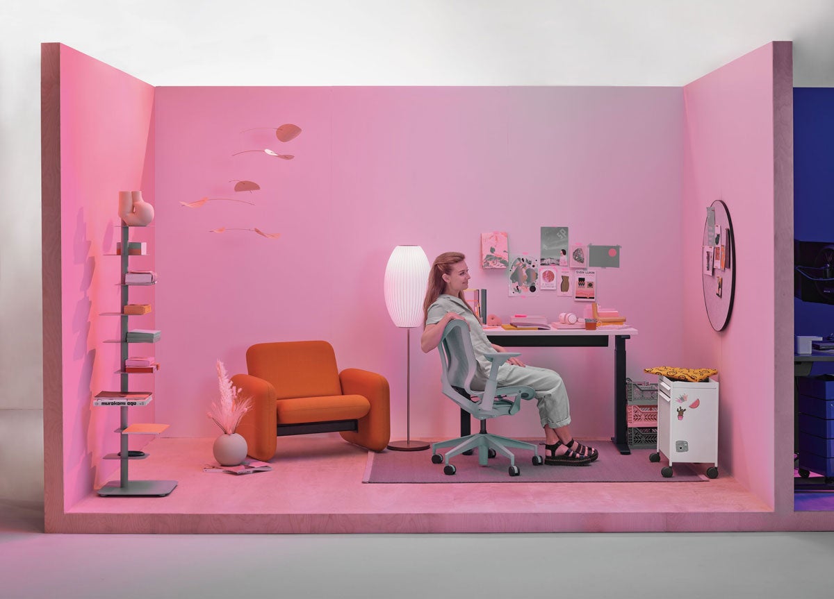 Thuiskantoor met vrouw zittend in een Cosm-bureaustoel met lage rugleuning in gletsjergrijs met een compact zit-sta-bureau en een Wilkes-fauteuil op de achtergrond