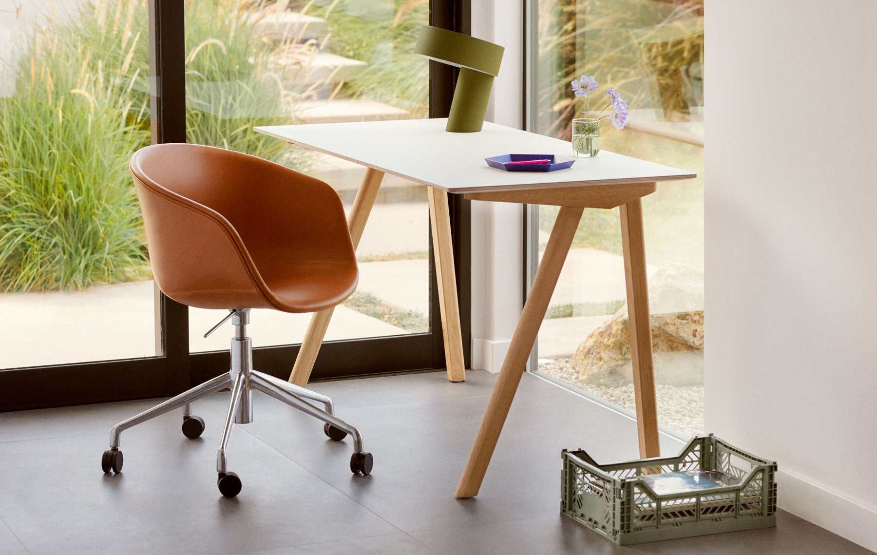 Eenvoudige foto in een huiselijke omgeving met uitzicht op de tuin, met een Hay Copenhague 90-bureau in eikenhout met een wit blad, een Hay Slant-lamp in kakigroen op het bureau en een kleine opbergkist van Hay op de vloer