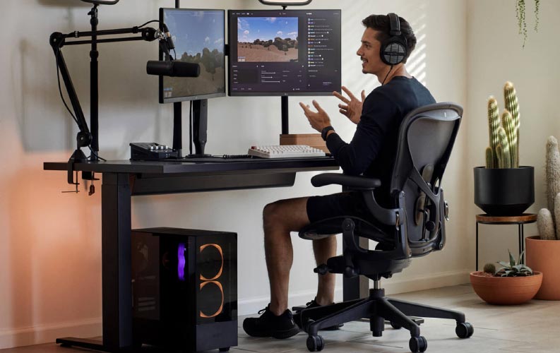 Gamer en streamer Xaryu in een gamingopstelling, zittend op een Herman Miller Aeron-gamingstoel met een zwart Nevi-sta- en zitbureau en twee monitoren op Ollin-monitorarmen