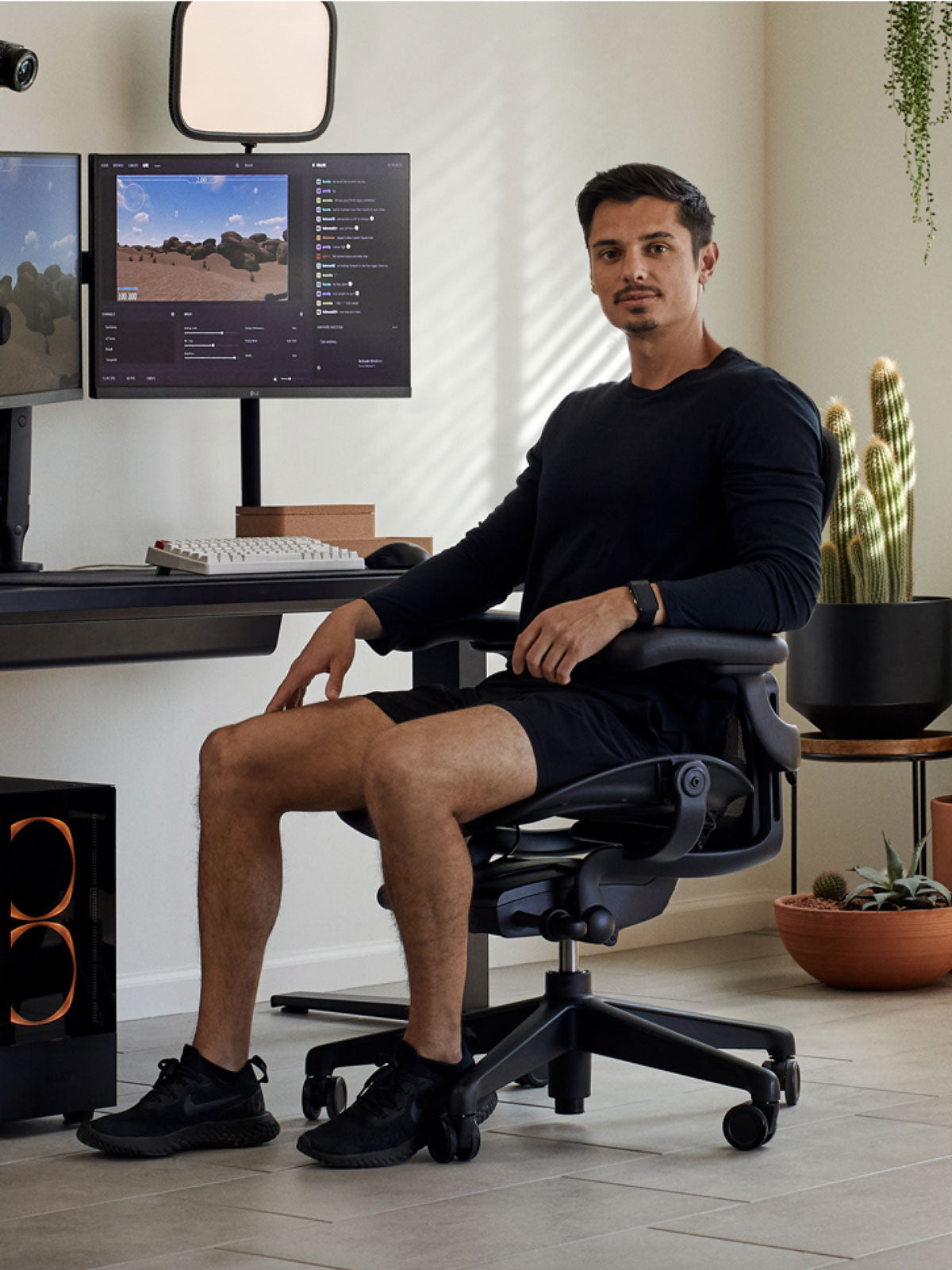 Een man zit in een zwarte Aeron-gamingstoel van Herman Miller, achter een zwart Nevi-bureau met een zwarte Ollin-monitorarm en -scherm. De foto is genomen in een binnenruimte met tegelvloeren, witte muren en kamerplanten in terracotta potten.