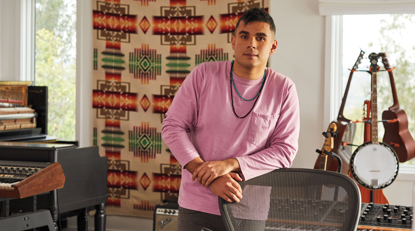 Rostam, muzikant, producer en componist, leunt tegen een Aeron-bureaustoel in grafiet met muziekinstrumenten op de achtergrond.